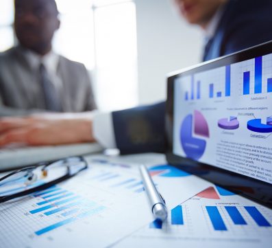 Image of eyeglasses, pen, touchpad and financial documents at workplace with businessmen working on background