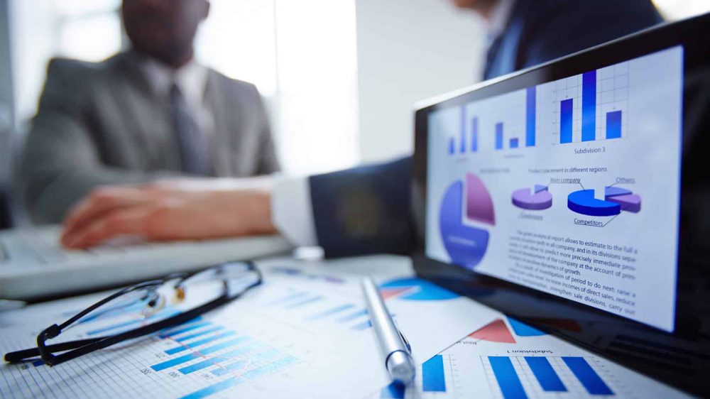 Image of eyeglasses, pen, touchpad and financial documents at workplace with businessmen working on background